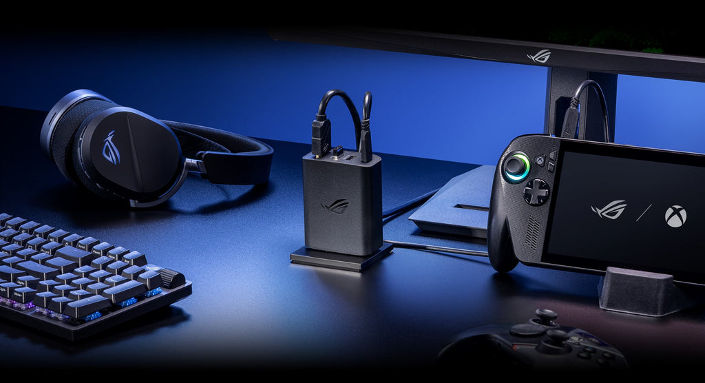 A close-up shot of the ROG 100W Gaming Charger Dock plugged into AC power in the center of a desk, with a monitor and ROG Xbox Ally X connected to the charger, with an ROG Raikiri Pro PC controller, an ROG wireless headset, and an ROG wireless keyboard visible in the foreground.