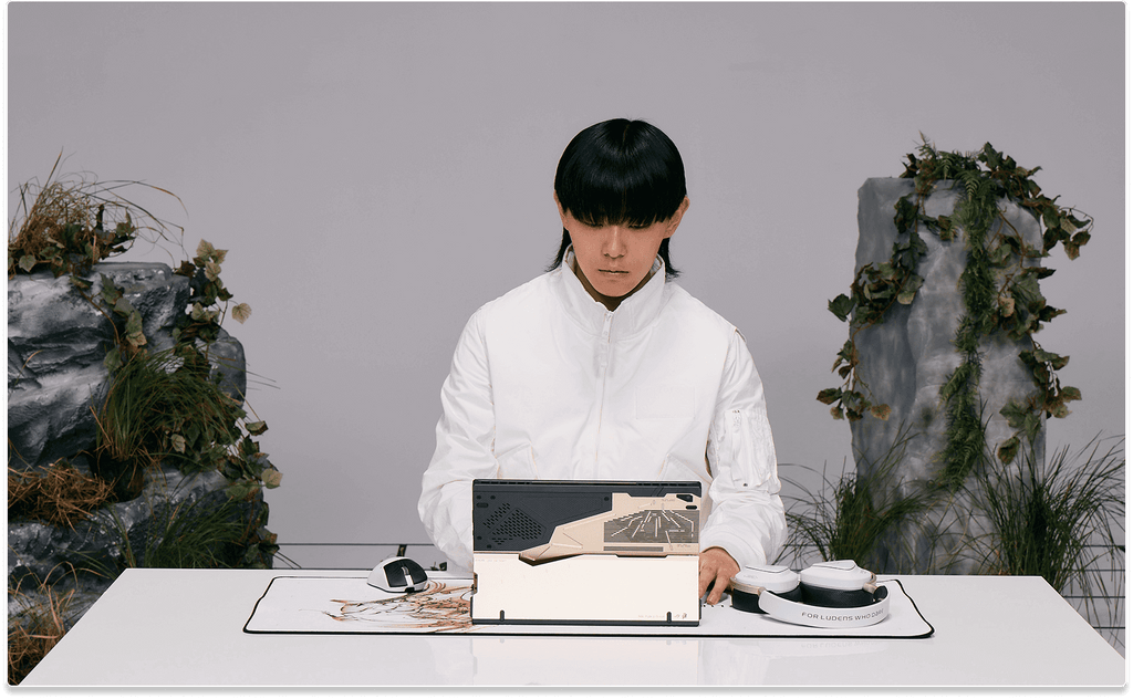 A man in white sitting at a desk typing on the Flow Z13 laptop, which is placed on the Scabbard II XXL KJP mouse pad and next to the Delta II KJP headset and a Keris II Origin KJP mouse