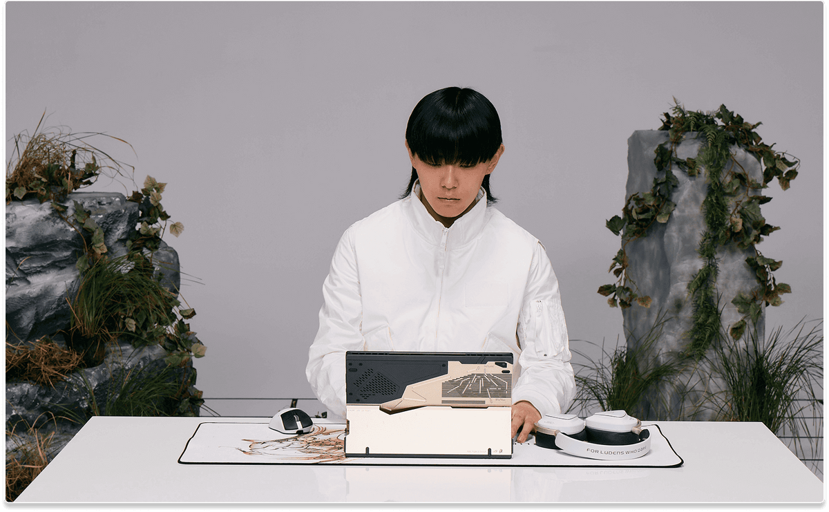 A man in white sitting at a desk typing on the Flow Z13 laptop, which is placed on the Scabbard II XXL KJP mouse pad and next to the Delta II KJP headset and a Keris II Origin KJP mouse