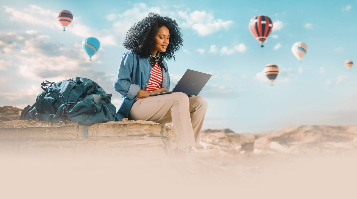 A young traveler seated outdoors on a rock, using a laptop with the RT-AX50 Go by side, against a backdrop of numerous hot air balloons dotting the blue sky.
