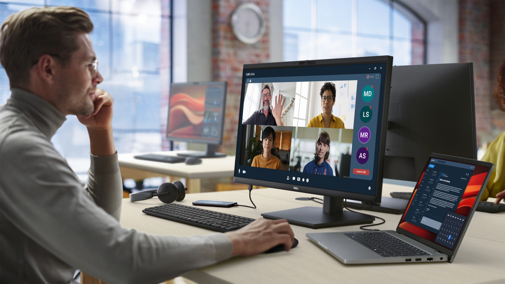 A man in an office uses a Dell Pro 24 Video Conferencing Monitor, along with a laptop, keyboard, mouse, and headset, to video chat with people.