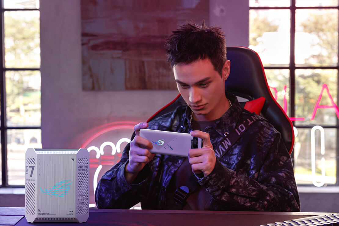 A man sitting in a gaming chair playing on an ROG phone, with the ROG Strix GS-BE7200 WiFi 7 router placed on the table in front of him in a daylight-lit gaming room.