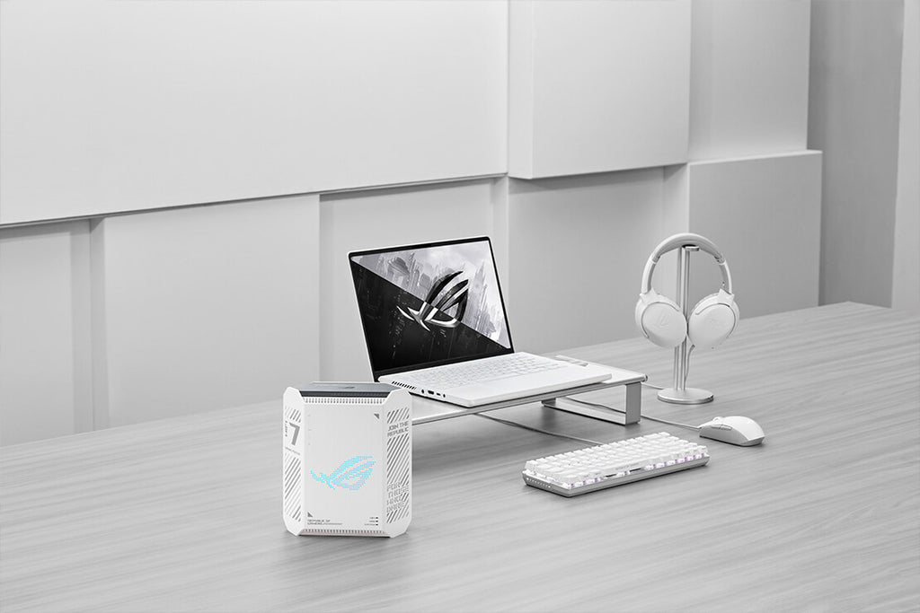 A minimalist workspace setup featuring a white ROG laptop, wireless headset, keyboard, and mouse, with the ROG Strix GS-BE7200 WiFi 7 router placed on the left side of the desk.