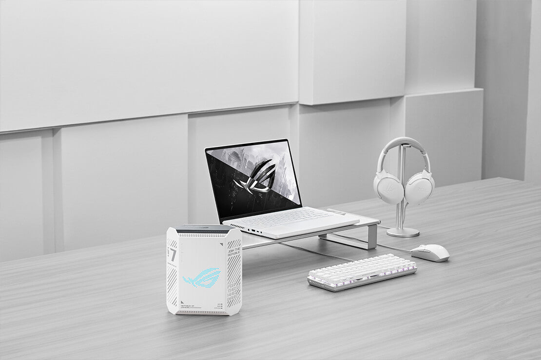 A minimalist workspace setup featuring a white ROG laptop, wireless headset, keyboard, and mouse, with the ROG Strix GS-BE7200 WiFi 7 router placed on the left side of the desk.