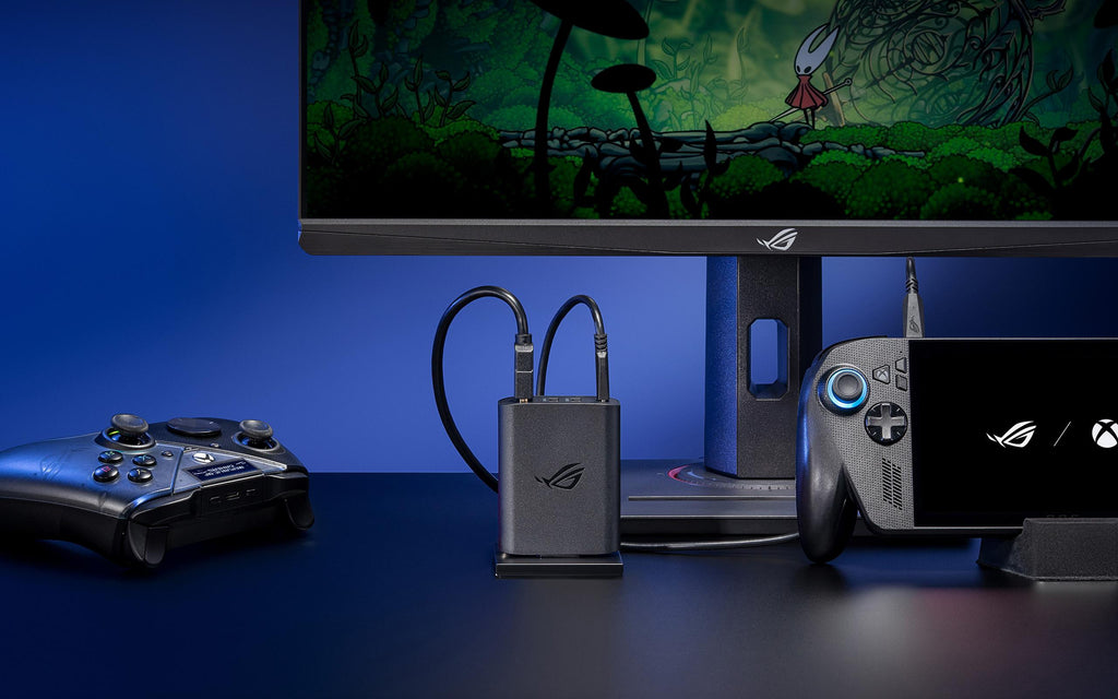 A close-up of an ROG 100W Gaming Charger Dock plugged into AC power in the center of a black desk, also plugged into a monitor and the ROG Xbox Ally X, with an ROG Raikiri Pro PC Controller, ROG Strix SCAR 16 laptop, and ROG wireless keyboard arranged in the foreground.