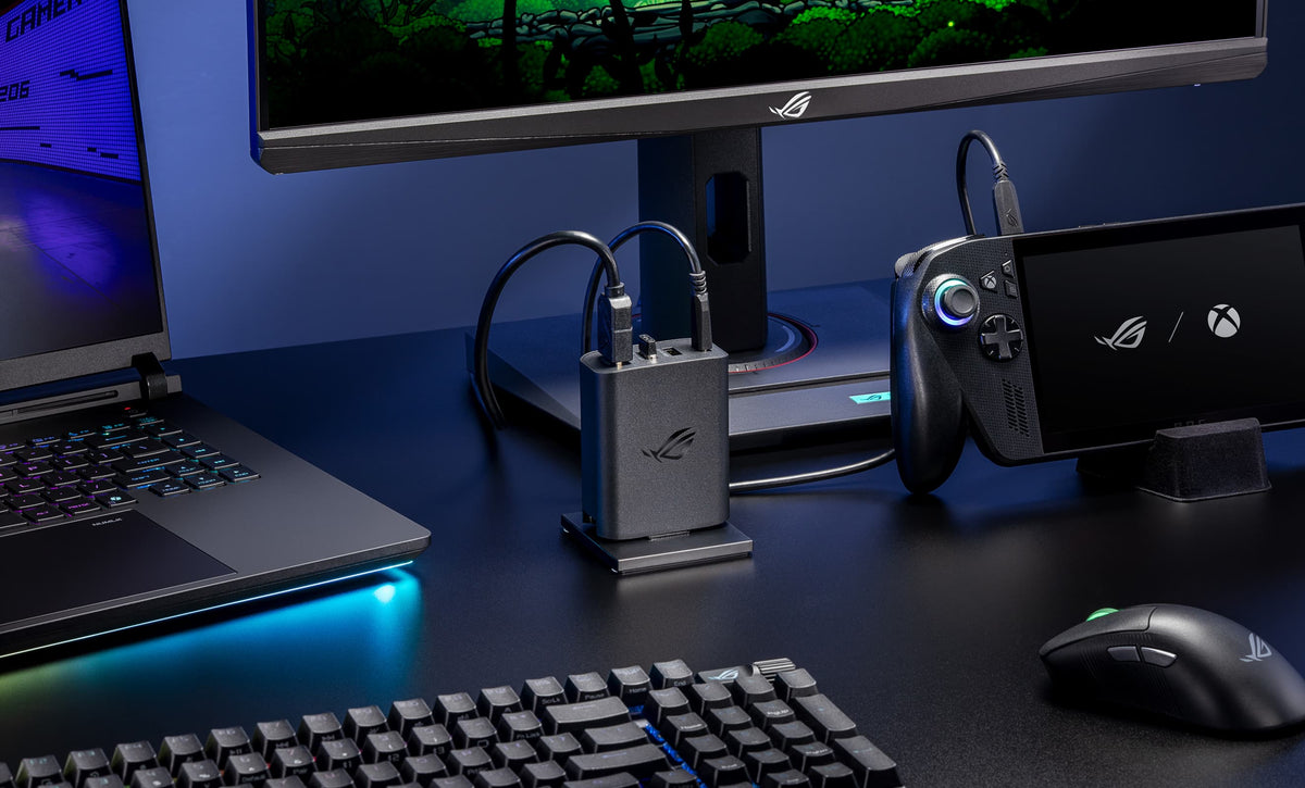 A close-up view of an ROG 100W Gaming Charger Dock plugged into AC power in the center of a desk, connected to a monitor and an ROG Xbox Ally X, with an ROG wireless mouse and keyboard, as well as an ROG Strix SCAR 16 laptop visible in the foreground.