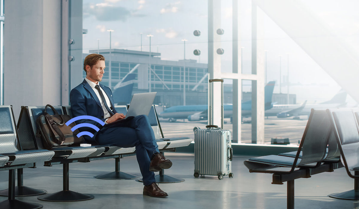 A businessman with the RT-AX50 Go router and suitcase by his side, sits in the airport using laptop, waiting for his flight.